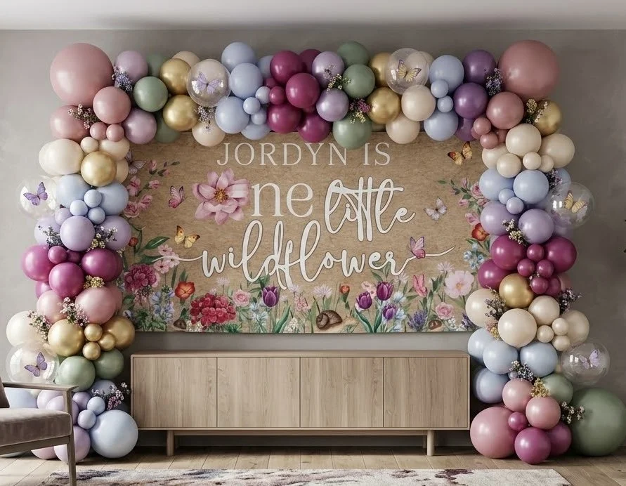 A pastel balloon arch in lilac, sage, blush and gold framing a hand-painted “one little wildflower” first-birthday sign above a console table