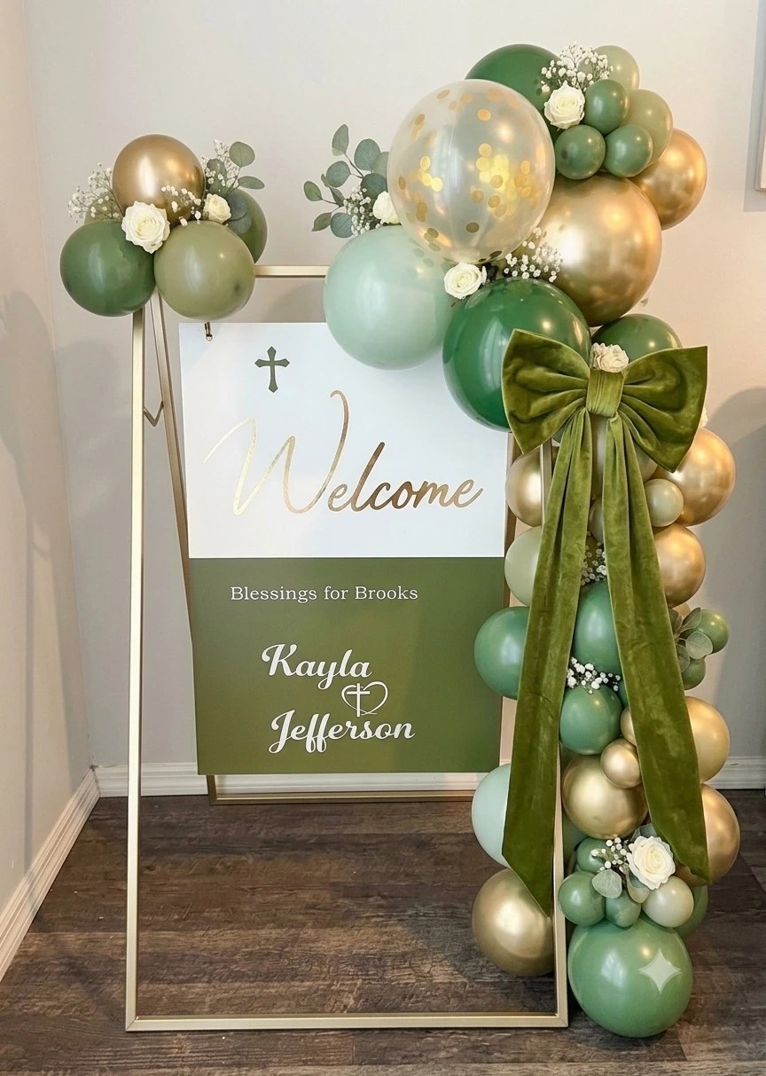 A sage, olive and gold balloon garland with white roses and a green velvet bow framing a gold-easel “blessings for Brooks” baptism welcome sign