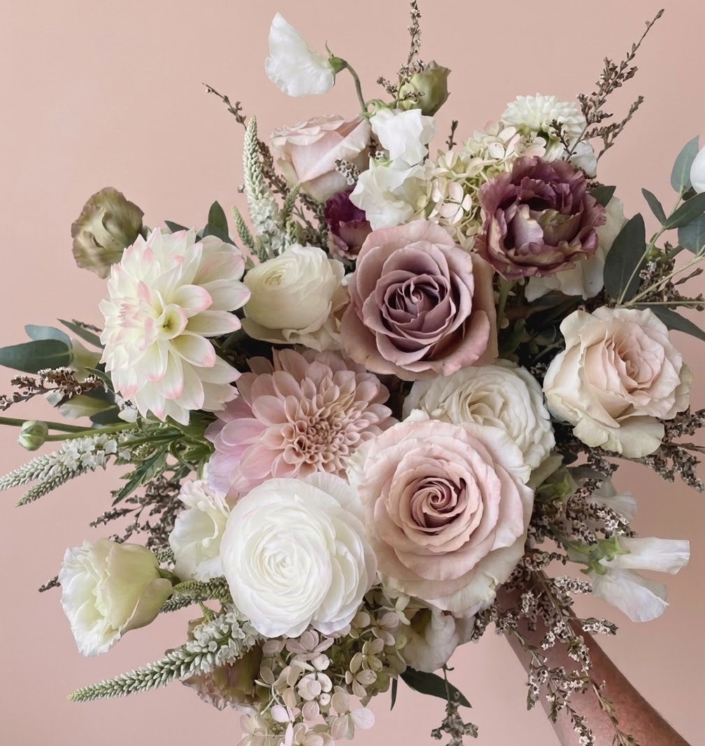 A hand-tied bouquet of mauve and blush roses, cream ranunculus, blush dahlias, sweet pea and eucalyptus against a pink wall