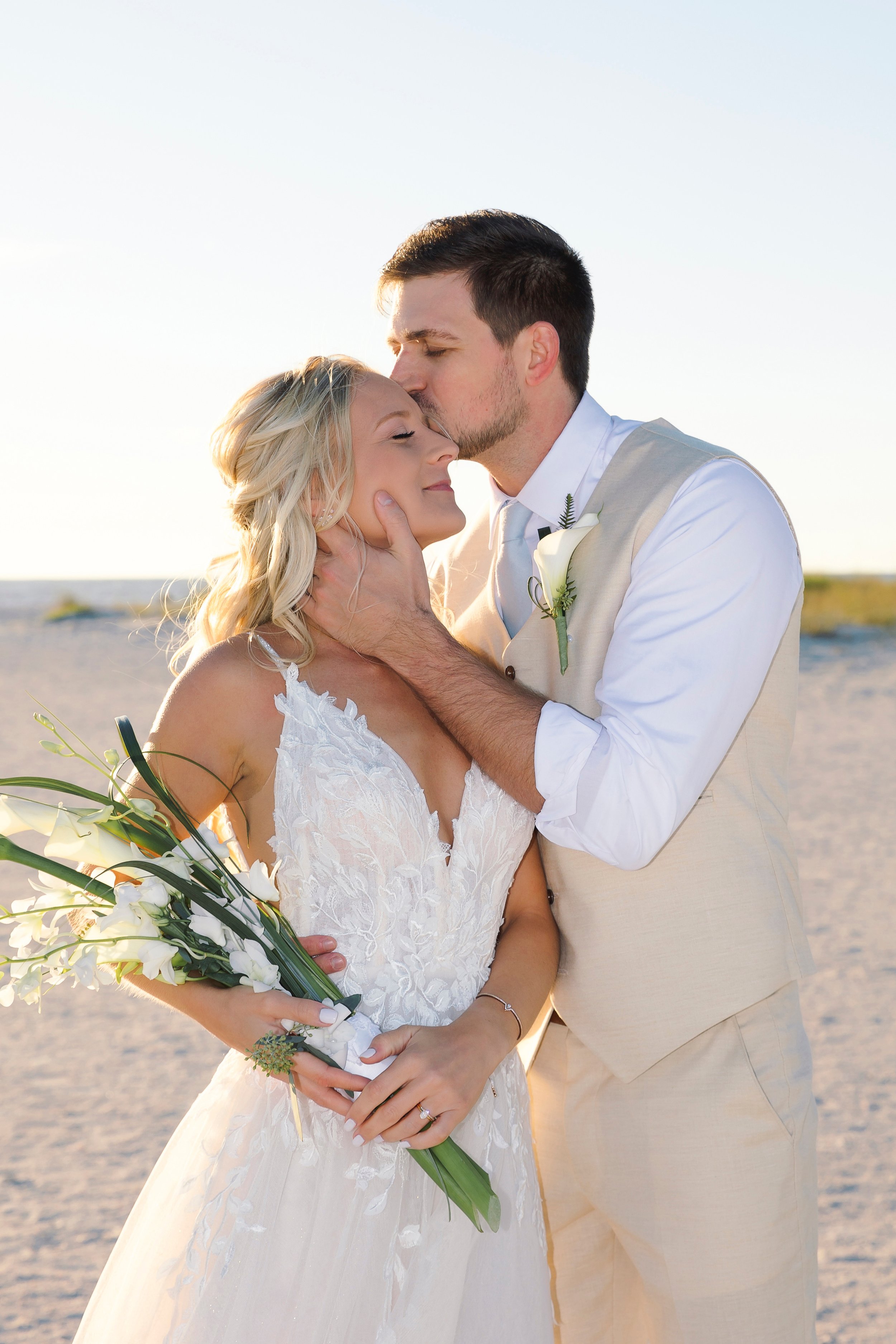 A groom kisses his bride's forehead on the beach; she carries a hand-tied bouquet of white blooms and greenery