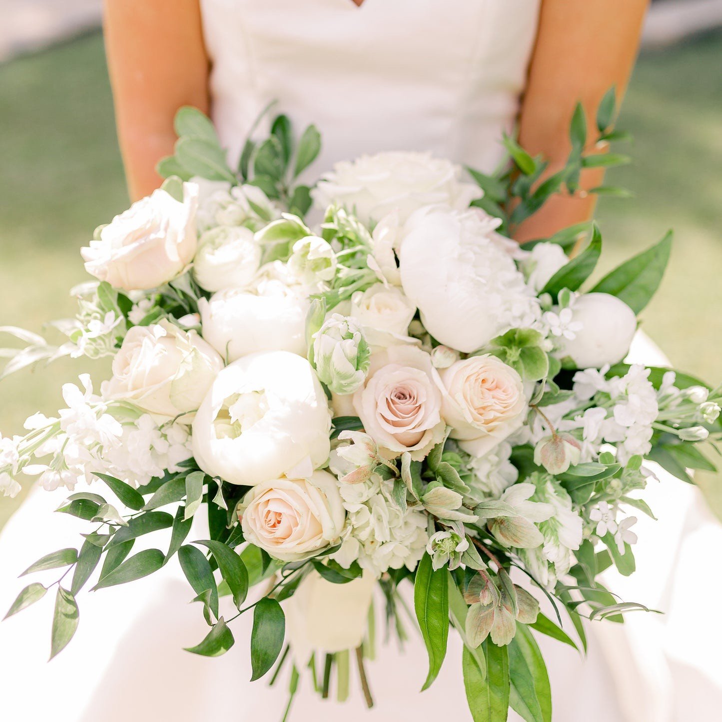 A bride holds a lush hand-tied bouquet of white peonies, blush garden roses and trailing greenery, designed by Julie