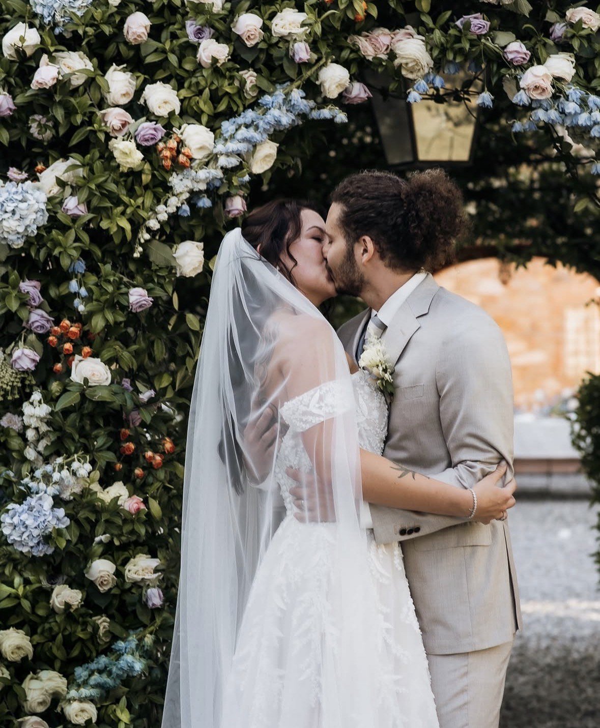A bride and groom share their first kiss beneath an arch of white, lilac and blue roses designed by Julie